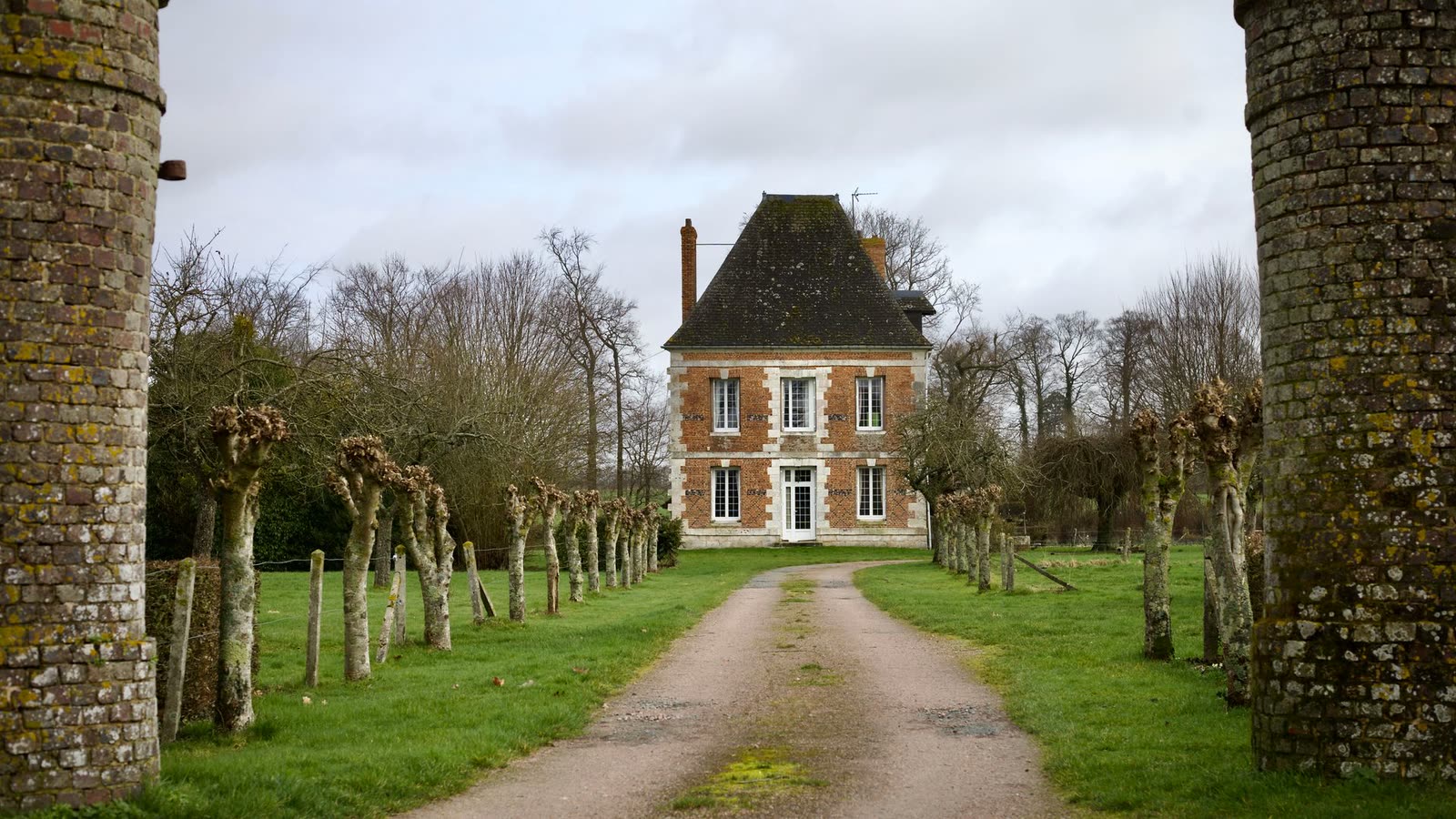 Maison de brique au bout d’une allée bordée d’arbres taillés